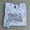 Folded gray sweatshirt with 'USA' text and flag on a gray surface