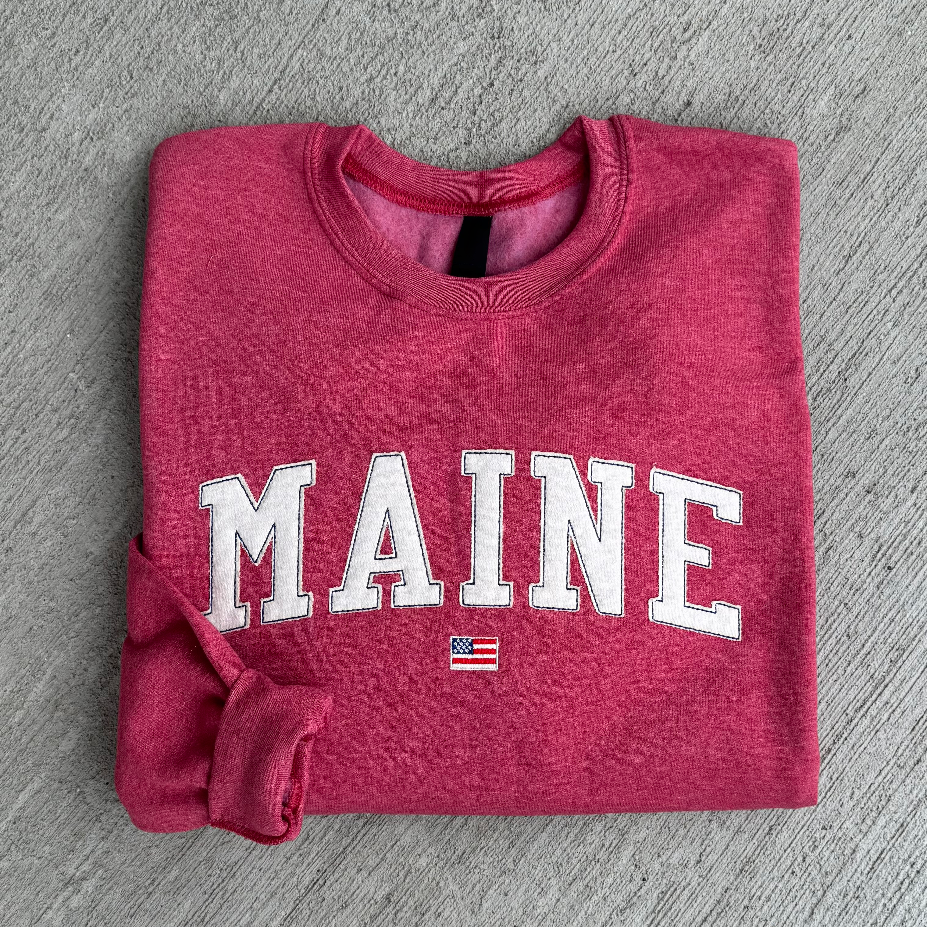Folded red sweatshirt with 'MAINE' fabric embroidered on it on a gray surface 