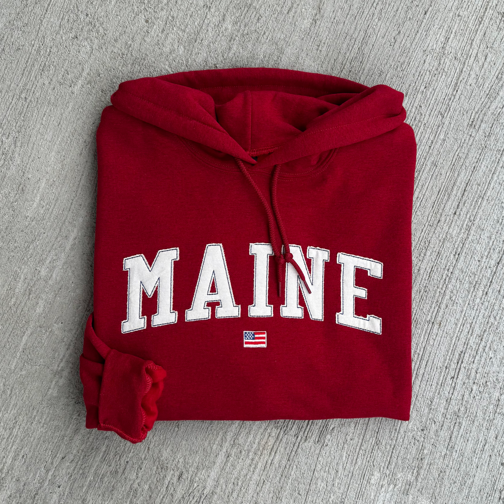 Folded red hoodie with 'MAINE' fabric embroidered on it against a gray background