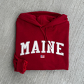 Folded red hoodie with 'MAINE' fabric embroidered on it against a gray background