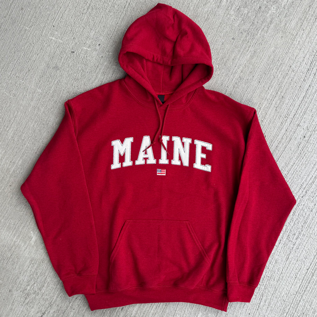 Red hoodie with 'MAINE' fabric embroidered on it, lying flat on a gray surface.