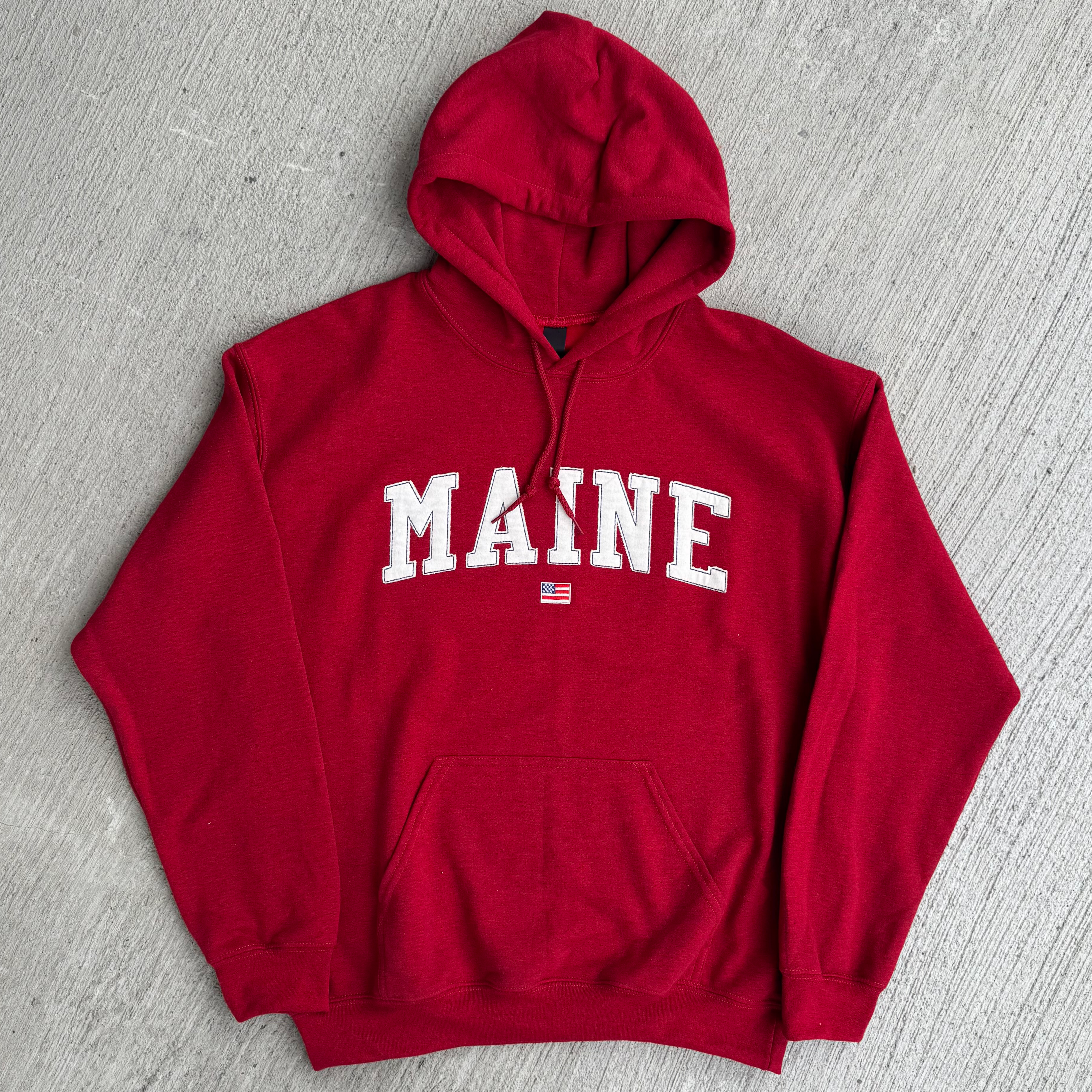 Red hoodie with 'MAINE' fabric embroidered on it, lying flat on a gray surface.