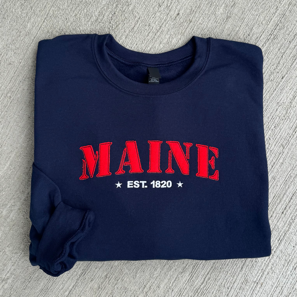 Folded navy blue sweatshirt with red 'MAINE EST. 1820' text on a gray surface