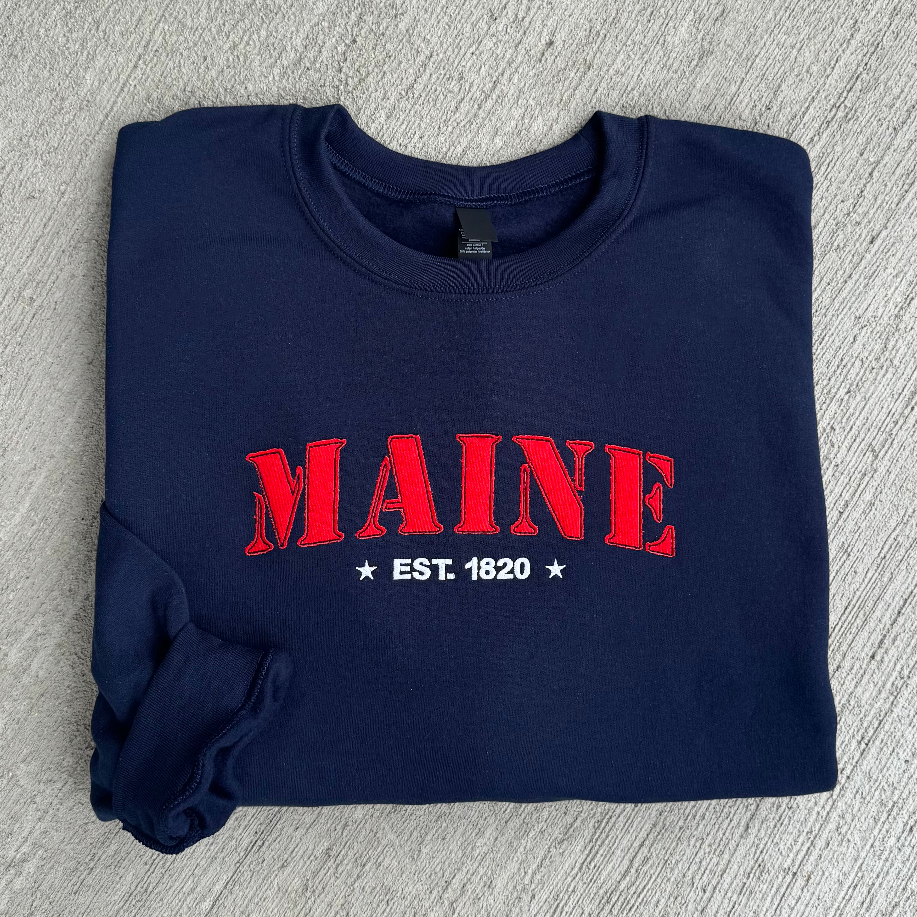 Folded navy blue sweatshirt with red 'MAINE EST. 1820' text on a gray surface