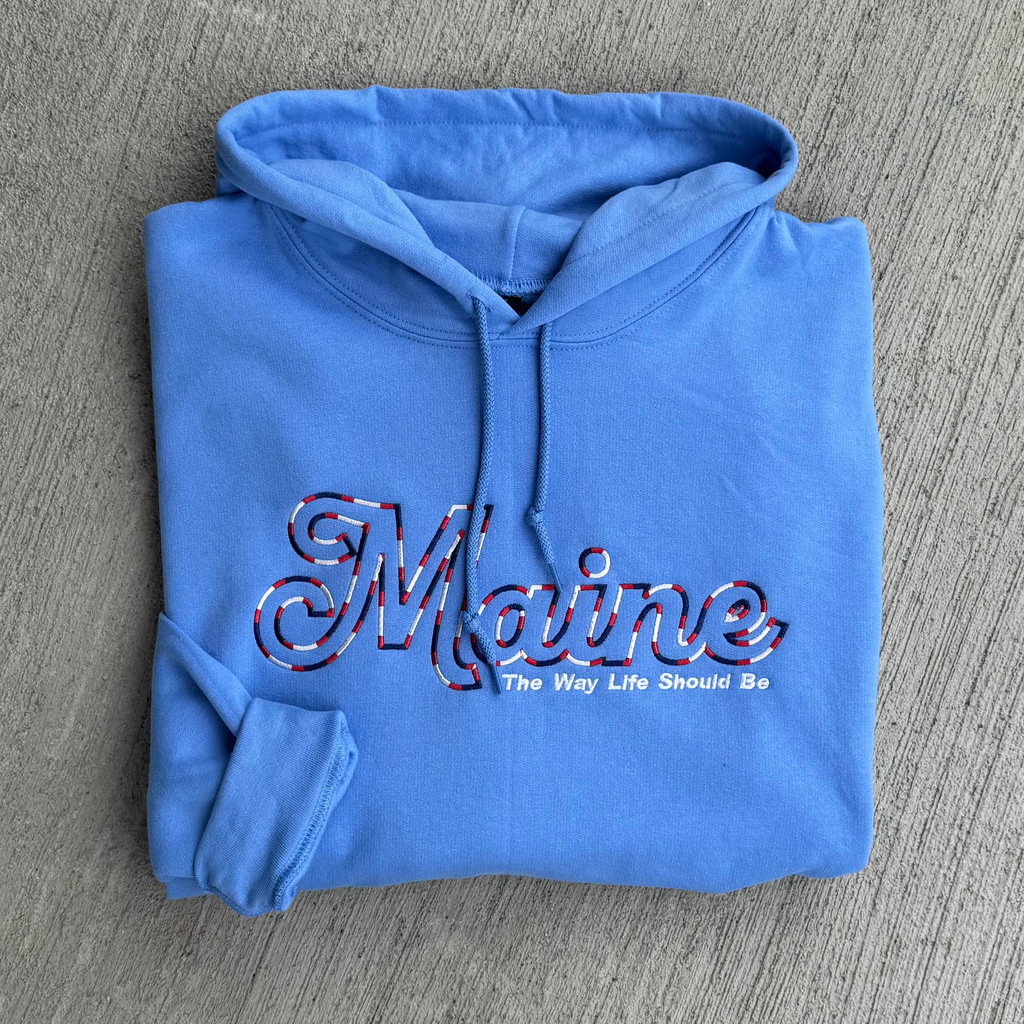 Folded blue hoodie with 'Maine The Way Life Should Be' text on a gray surface