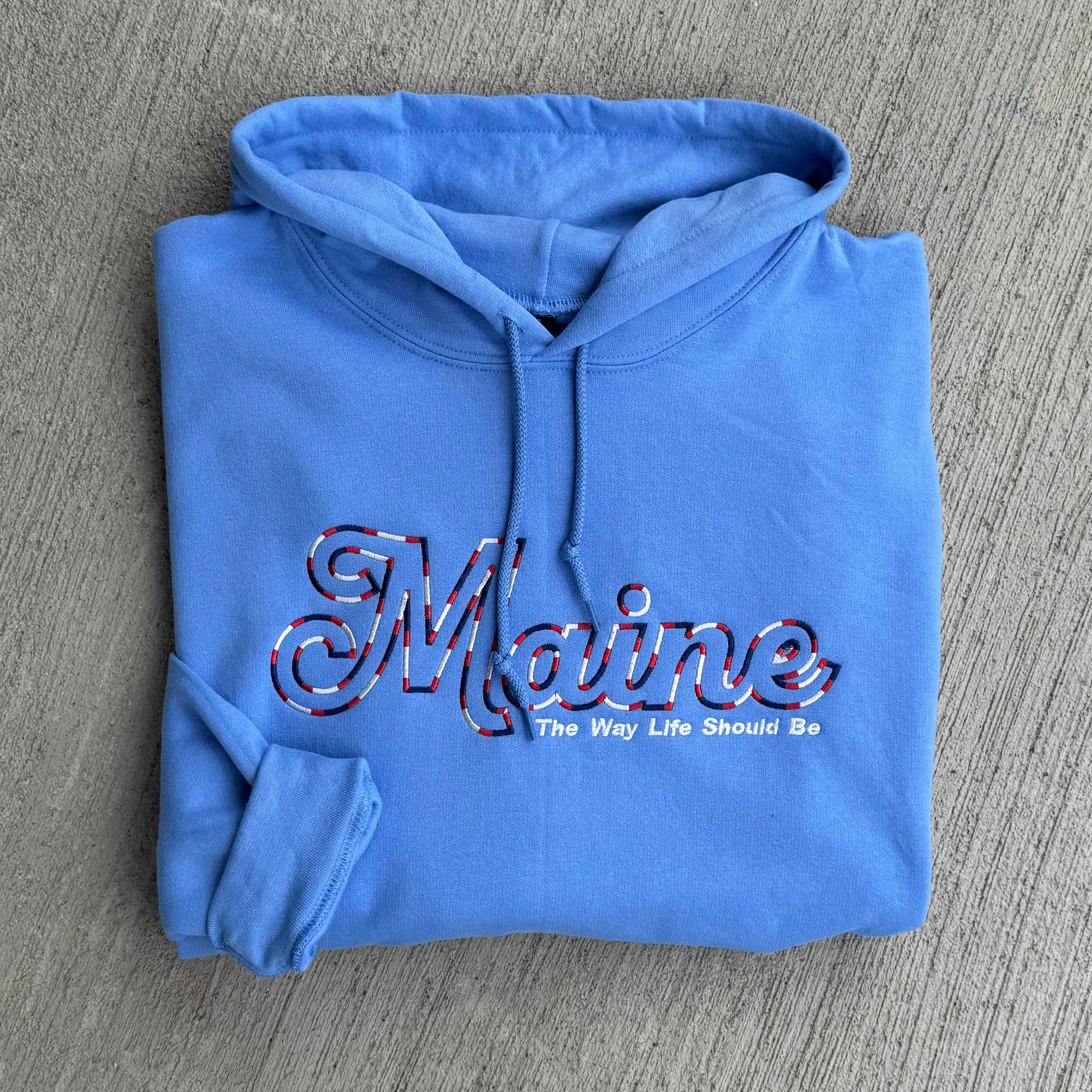Folded blue hoodie with 'Maine The Way Life Should Be' text on a gray surface