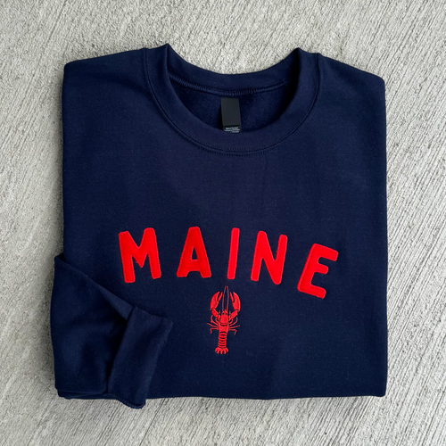 Folded navy blue sweatshirt with red 'MAINE' text and lobster design on a gray surface