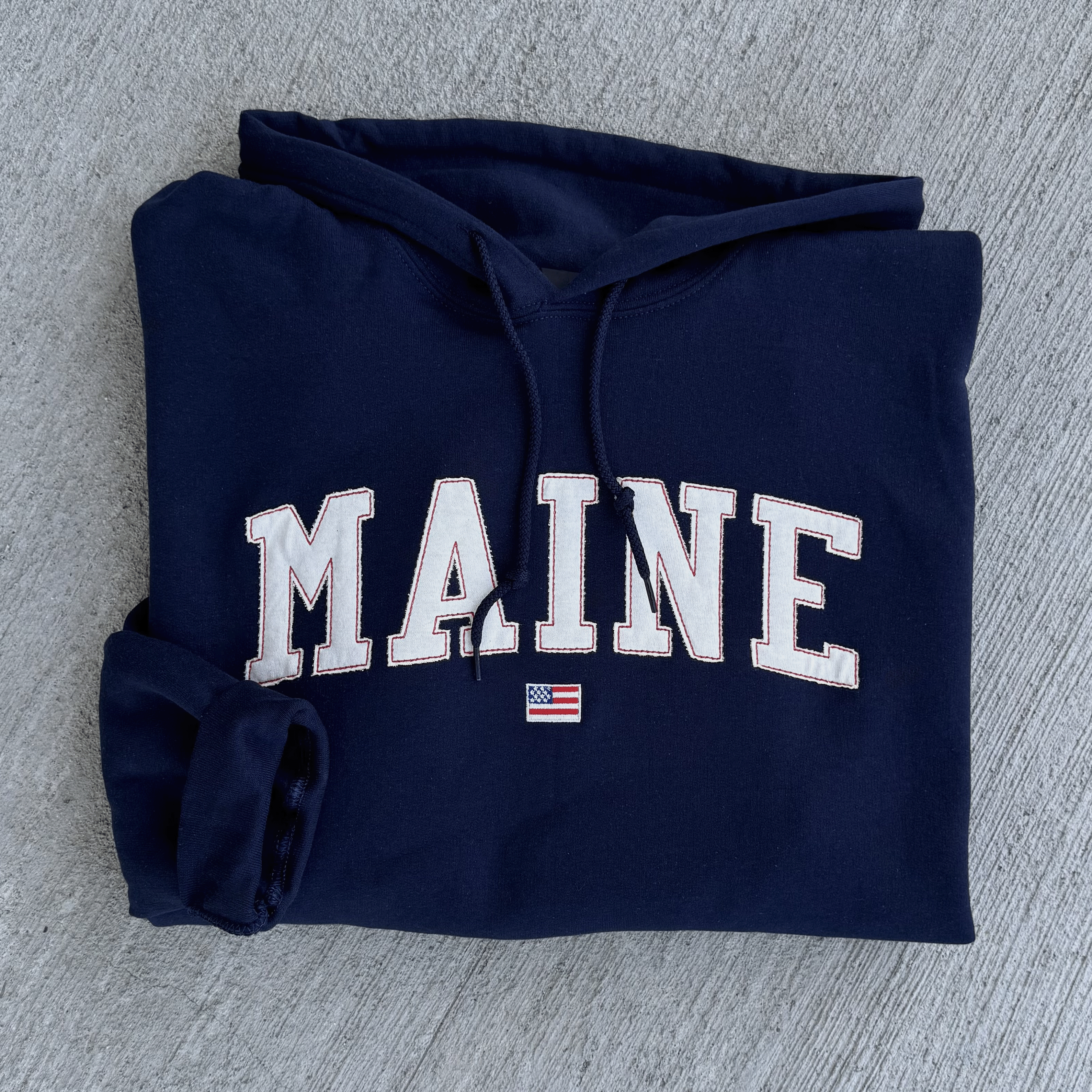 Folded navy blue hoodie with 'MAINE' text on a gray surface