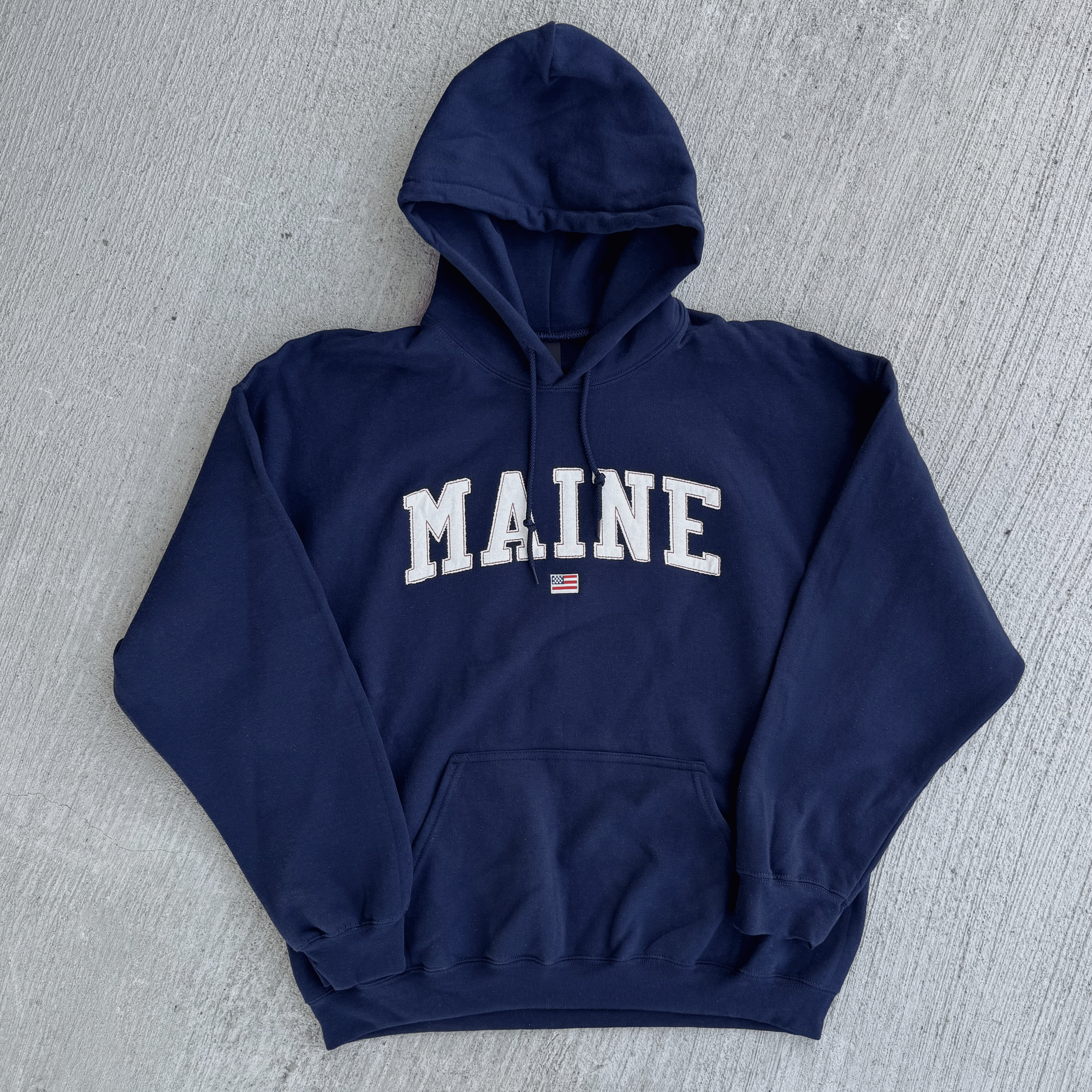 Navy blue hoodie with 'MAINE' printed on it, lying flat on a gray surface.