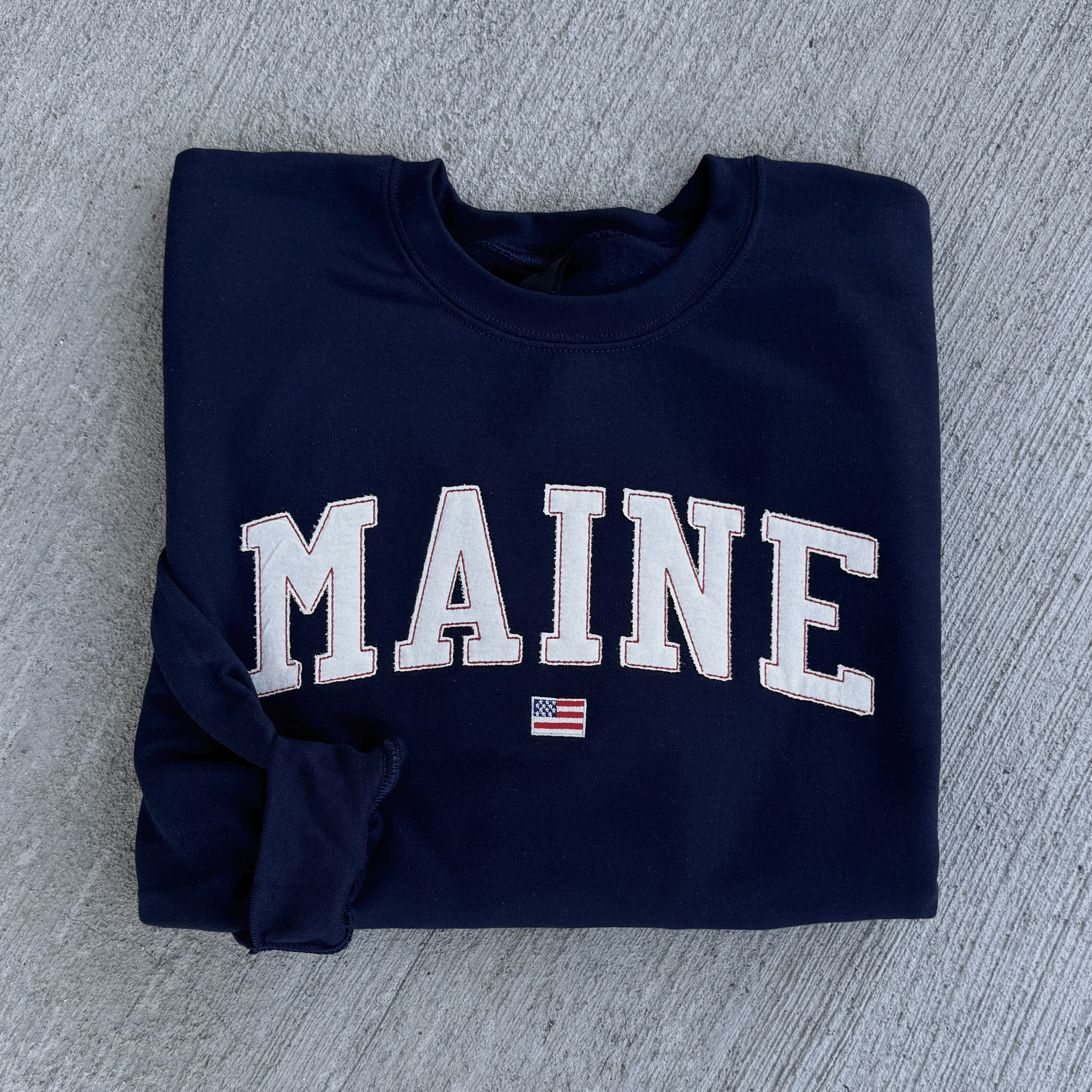 Folded navy blue sweatshirt with 'MAINE' printed on it, featuring an American flag, on a gray textured surface.