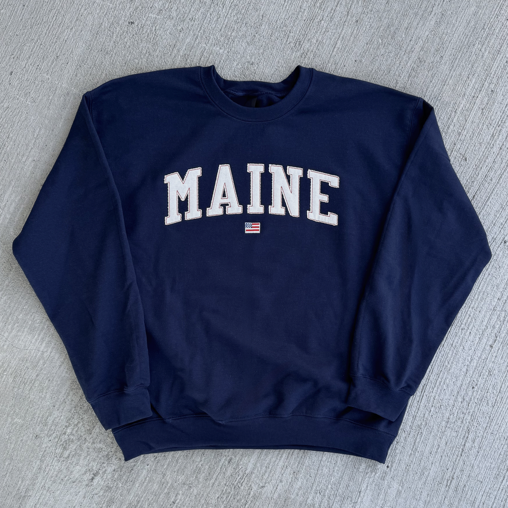 Navy blue sweatshirt with 'MAINE' printed on it, lying flat on a gray surface.