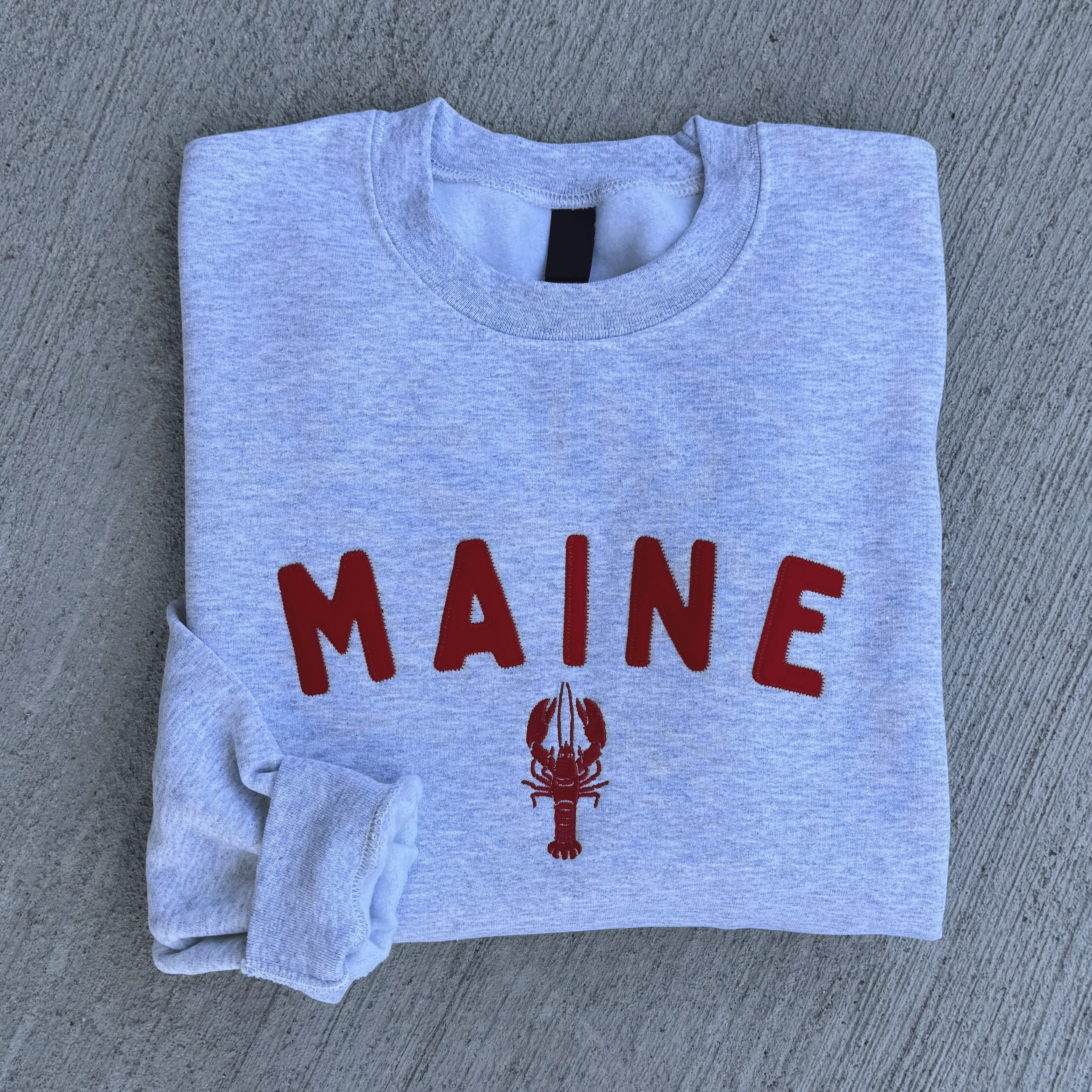 Folded gray sweatshirt with 'MAINE' and a lobster graphic on a gray surface