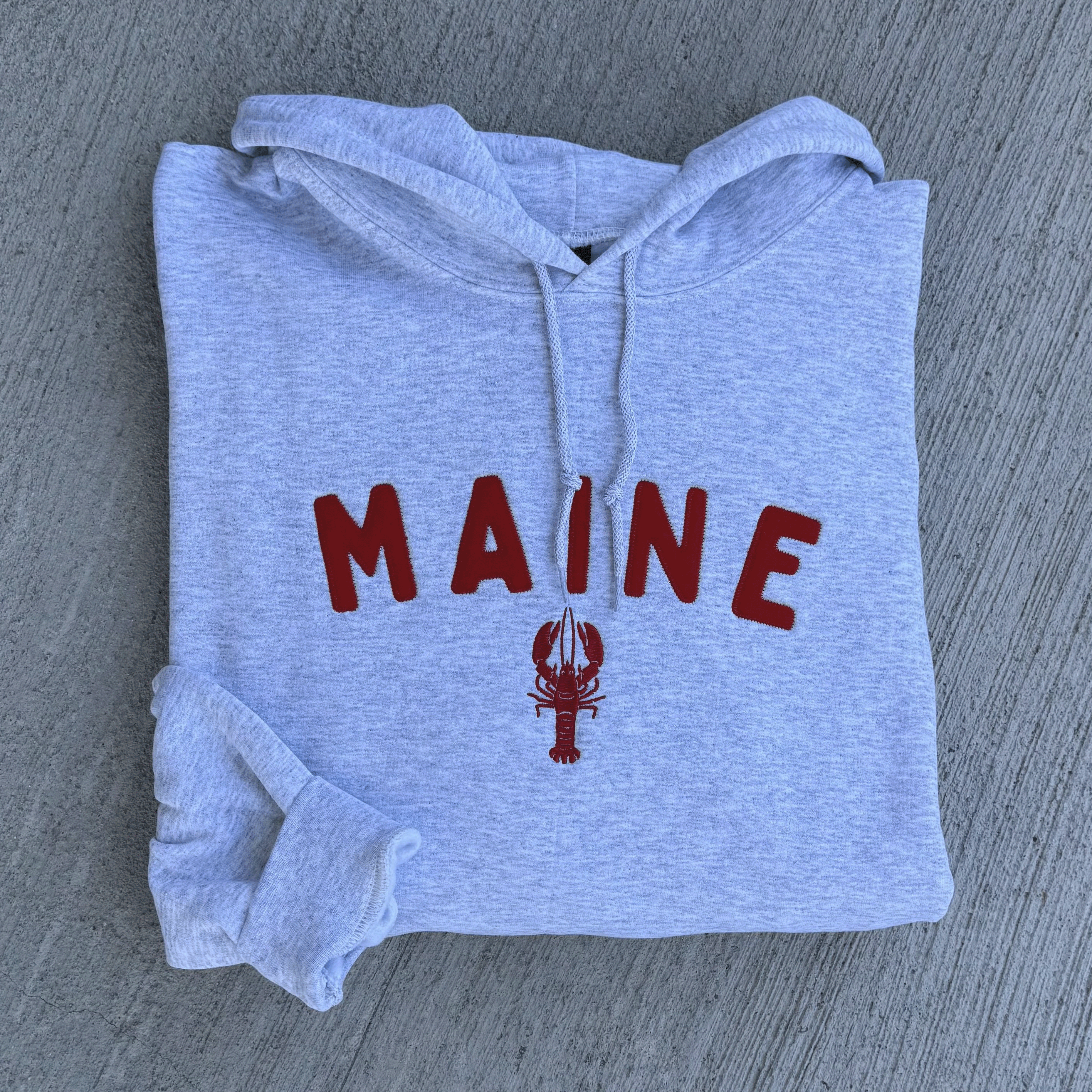 Folded gray hoodie with 'MAINE' and a lobster graphic on a textured surface