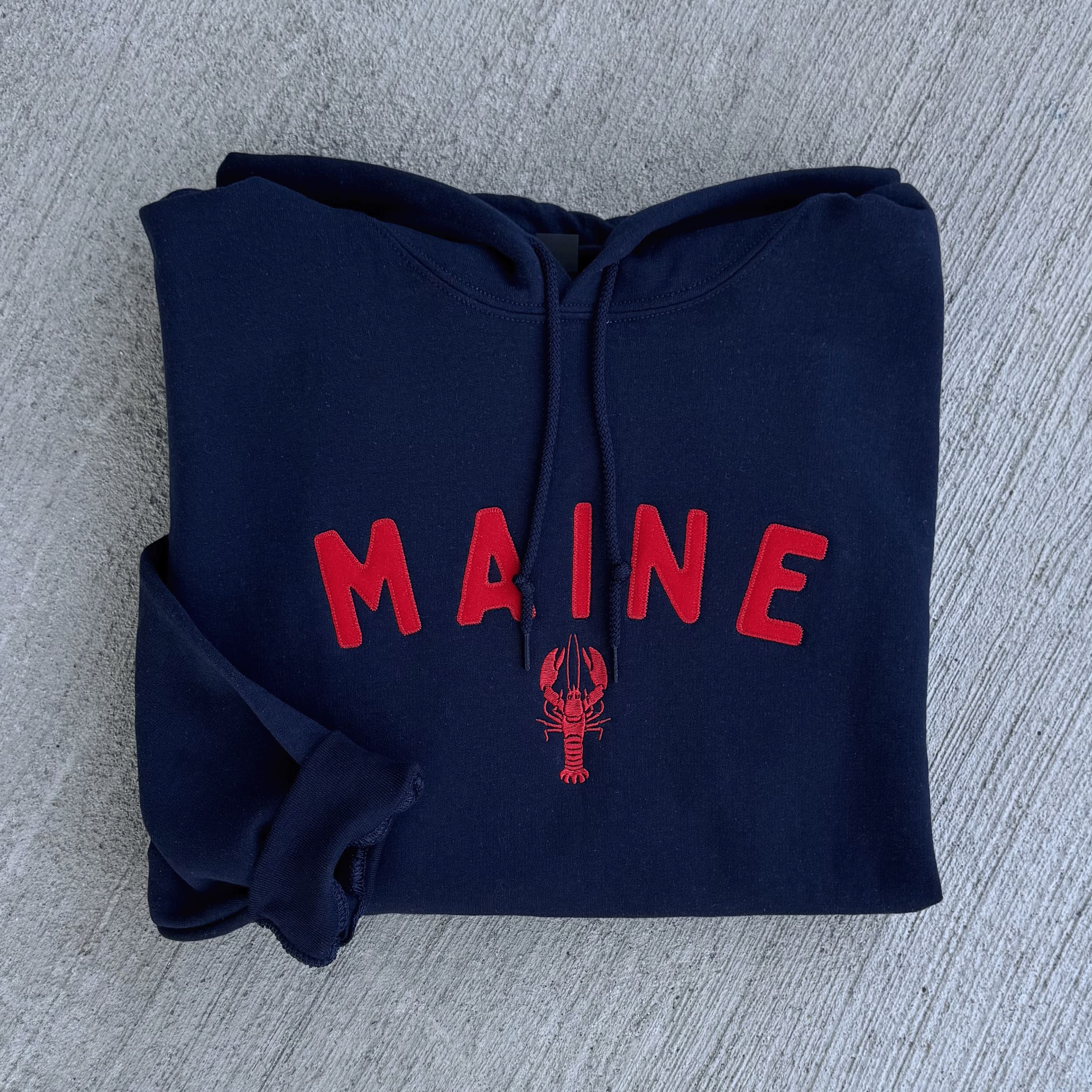 Folded navy blue hoodie with 'MAINE' in red text on a gray surface