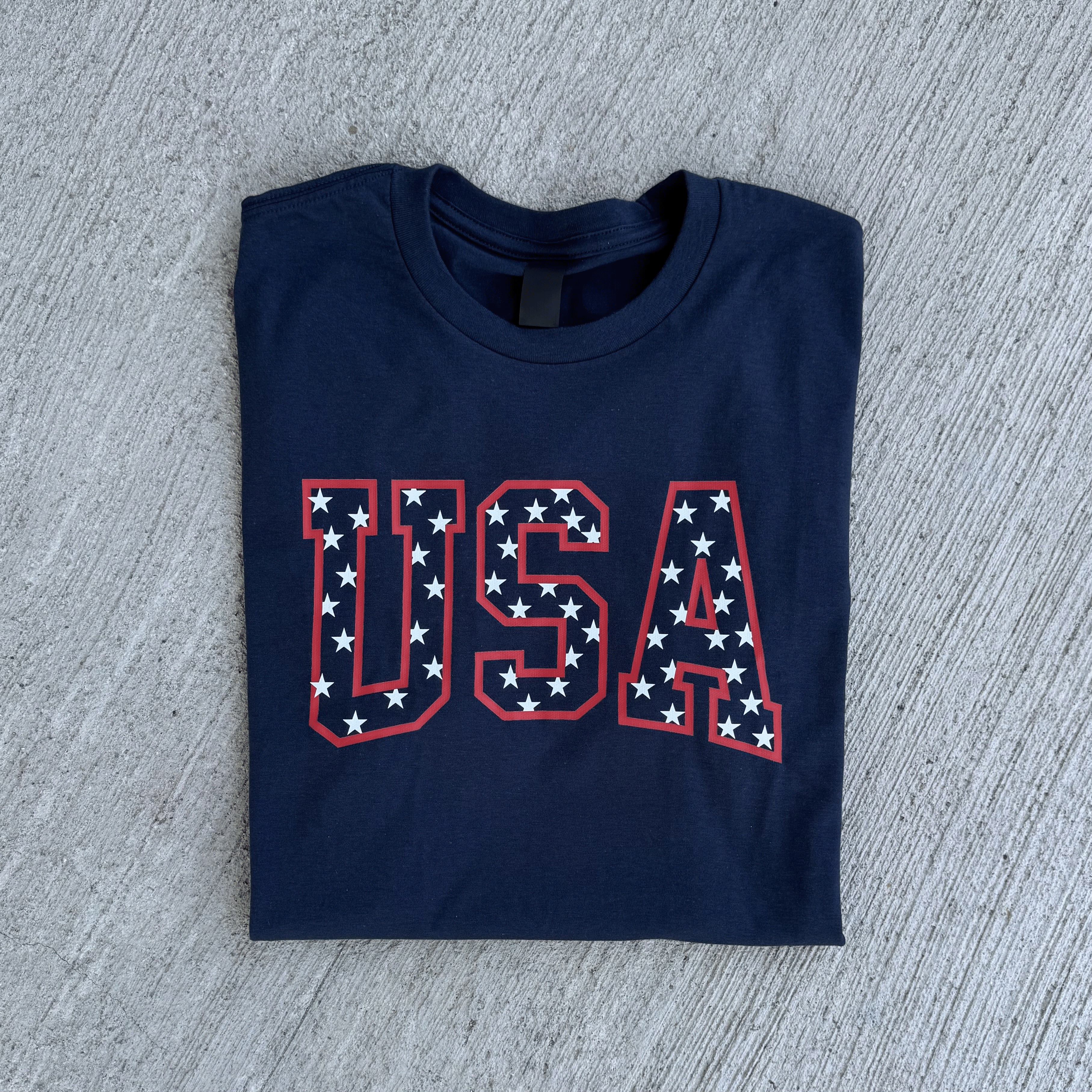 Folded navy blue t-shirt with 'USA' print on a gray concrete surface