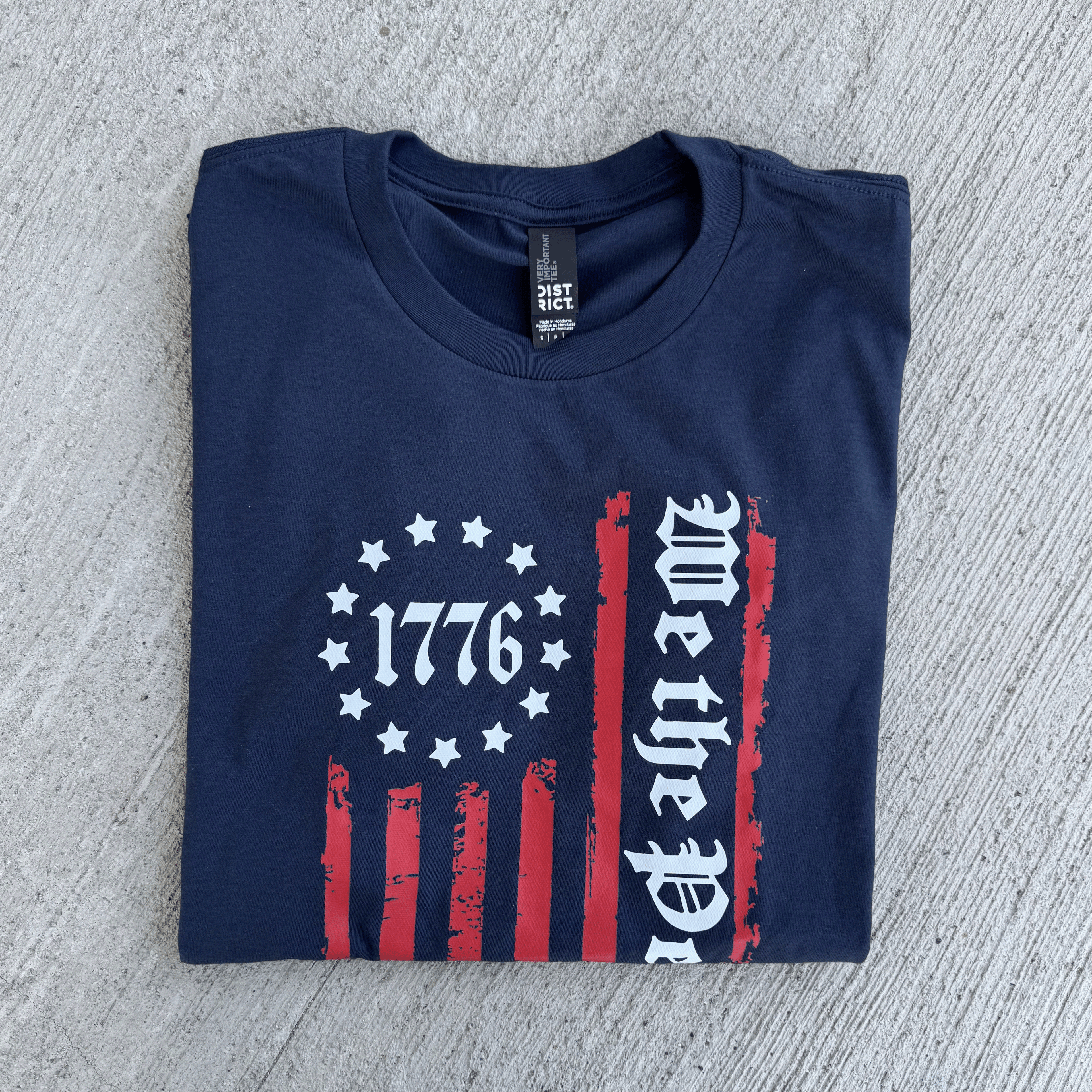 Folded navy blue t-shirt with 1776 and 'We the People' design on a textured surface