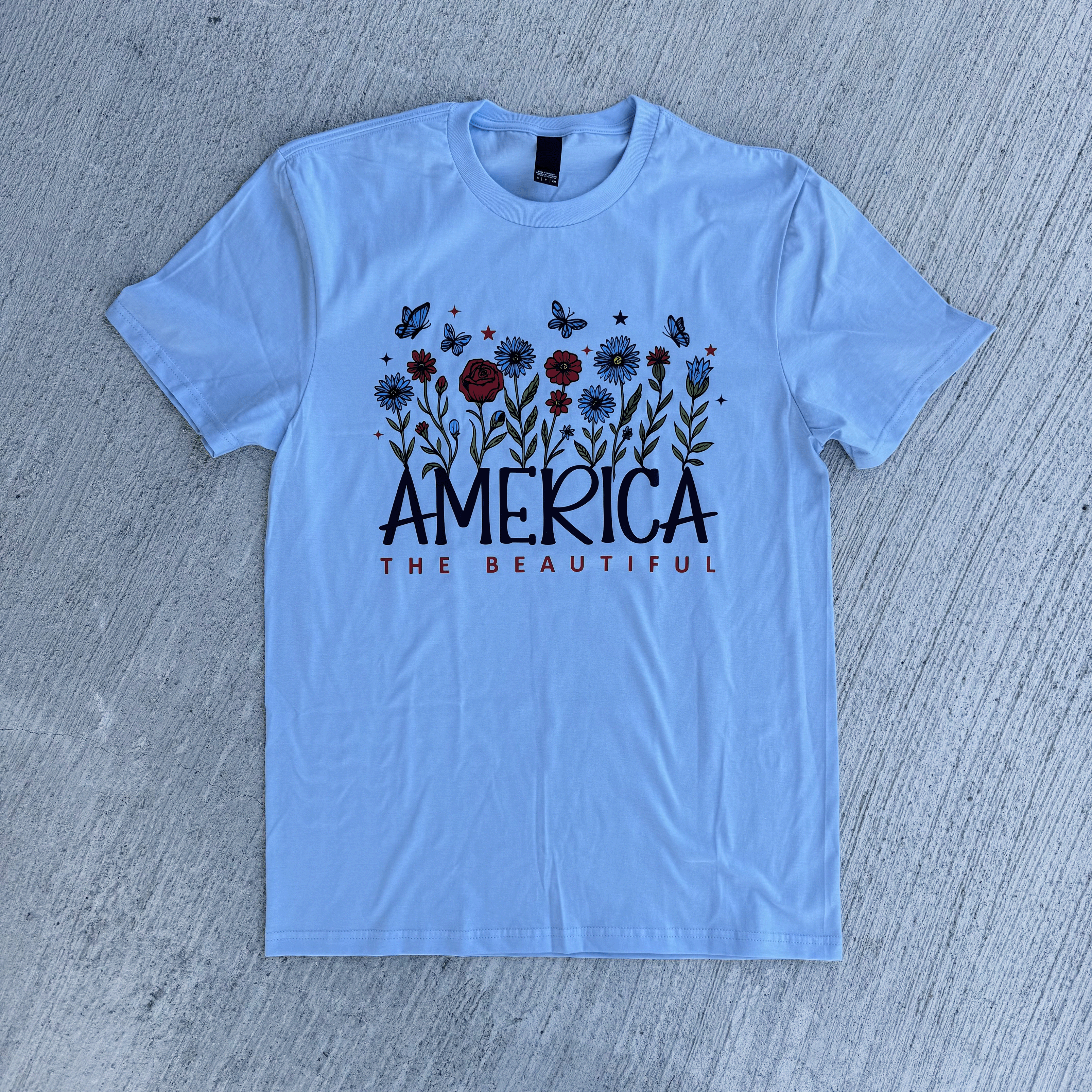 Light blue t-shirt with 'America the Beautiful' design on a gray surface