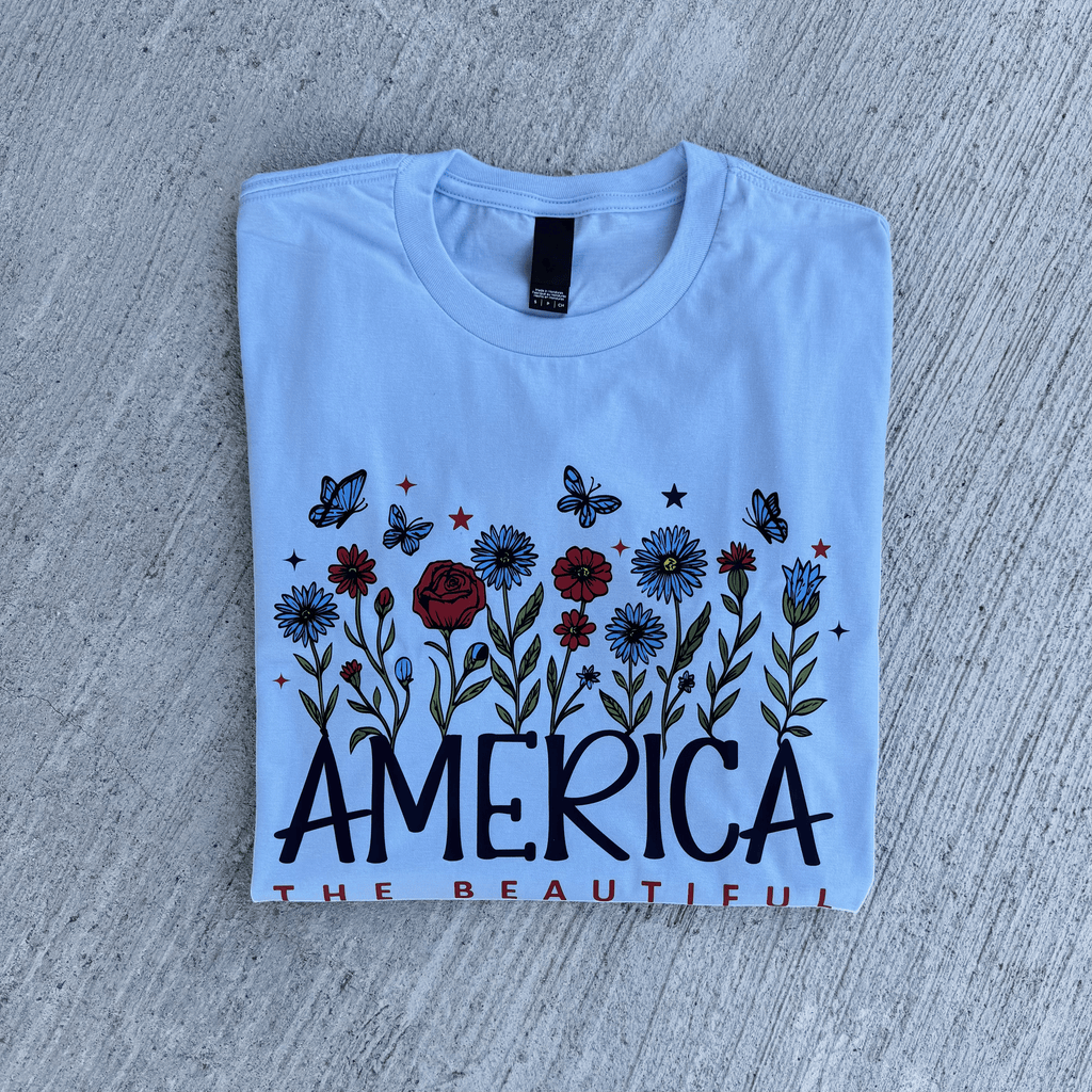 Folded light blue t-shirt with floral design and 'AMERICA THE BEAUTIFUL' text on a concrete surface.