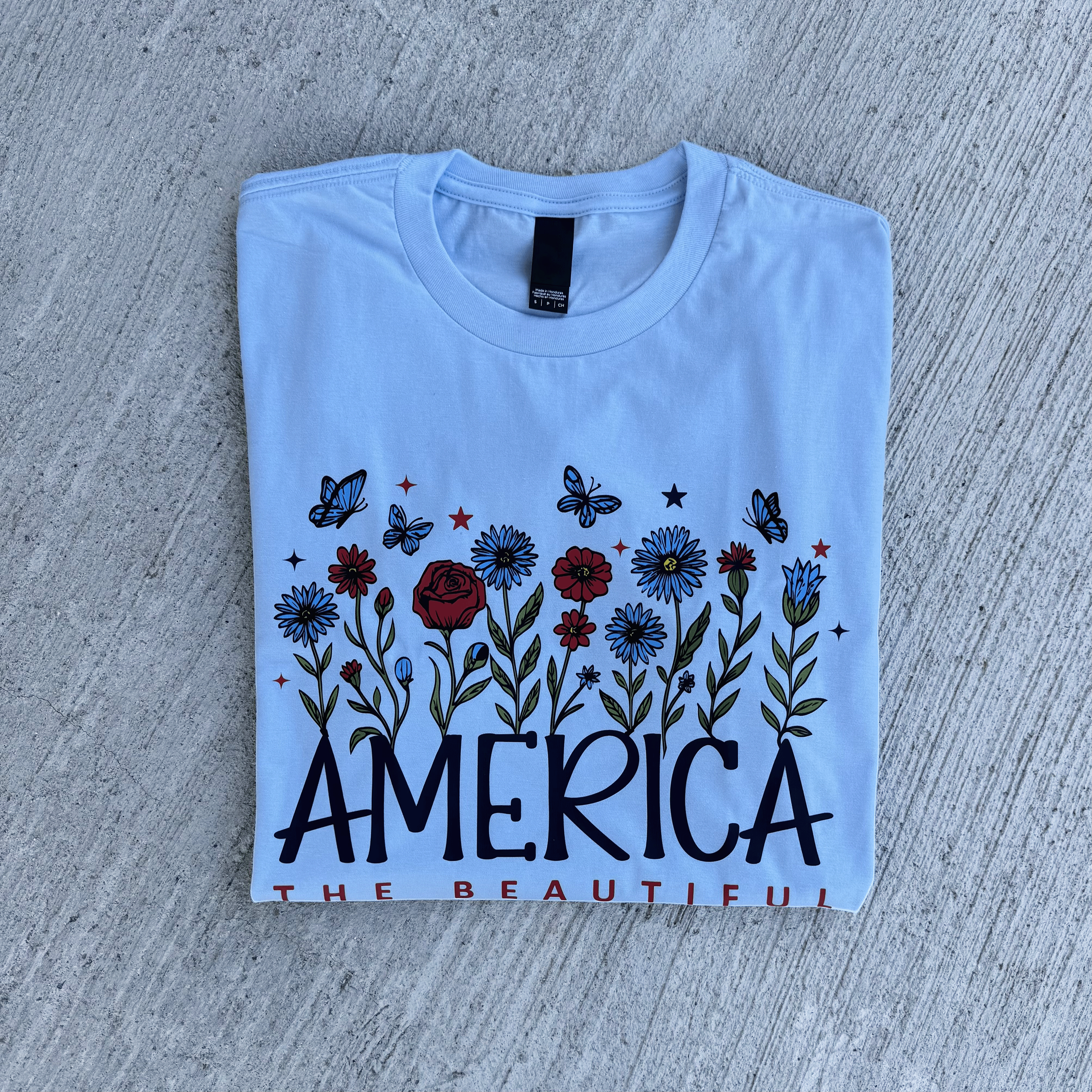 Folded light blue t-shirt with floral design and 'AMERICA THE BEAUTIFUL' text on a concrete surface.