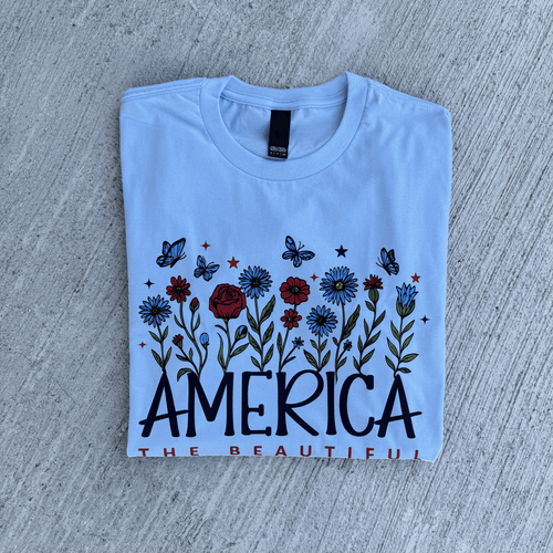 Folded light blue t-shirt with floral design and 'AMERICA THE BEAUTIFUL' text on a concrete surface.
